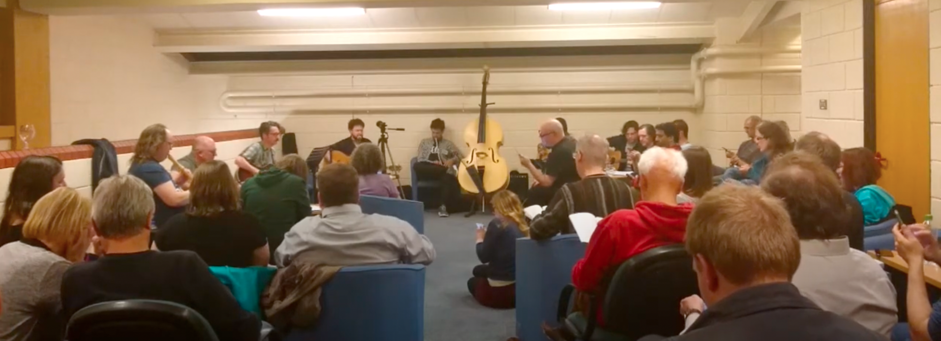 A large room is filled with people, mostly facing away from the camera towards the front of the space, where a selection of musicians are playing instruments. The people include a range of ages and genders, and a large double bass is visible in the centre