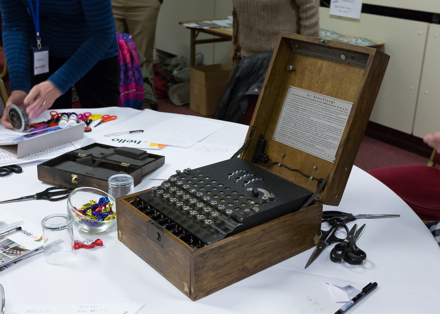 A Saturday night table. An Enigma machine sits on the table, surrounded by craft materials and paper. In the background, a person is holding a rotor from the Enigma machine.